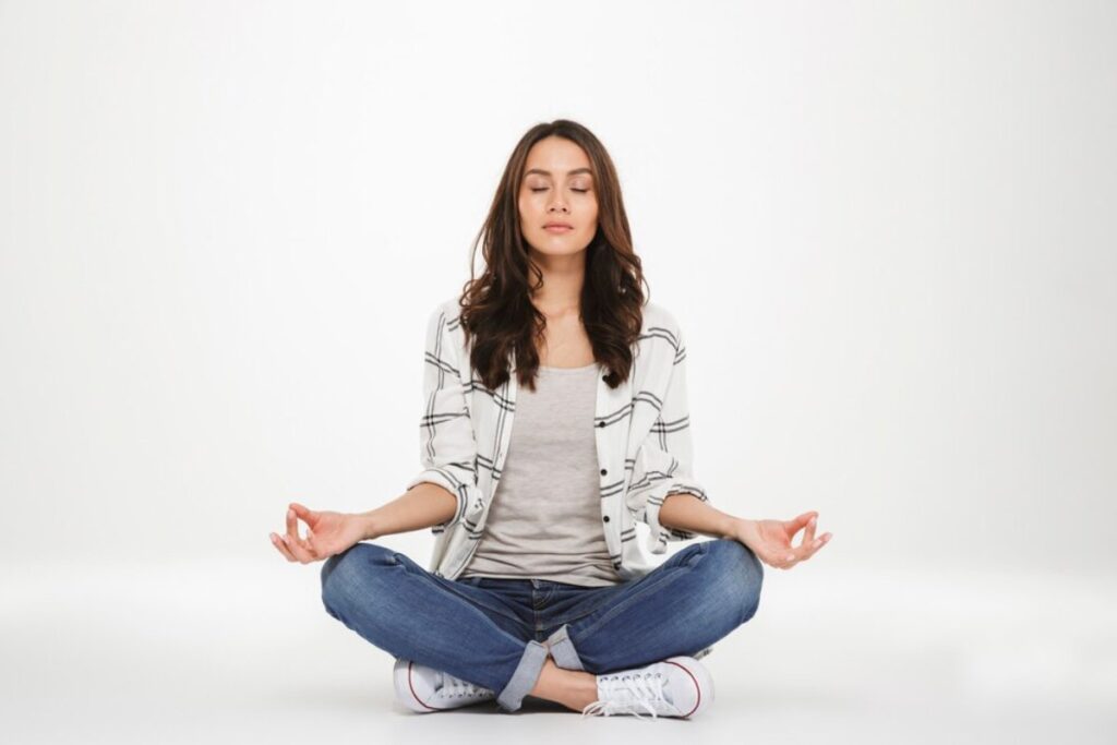 A woman sits cross-legged on a light background, practicing meditation, wearing a gray shirt and plaid jacket.