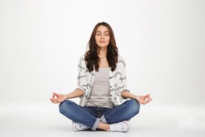 A woman sits cross-legged on a light background, practicing meditation, wearing a gray shirt and plaid jacket.