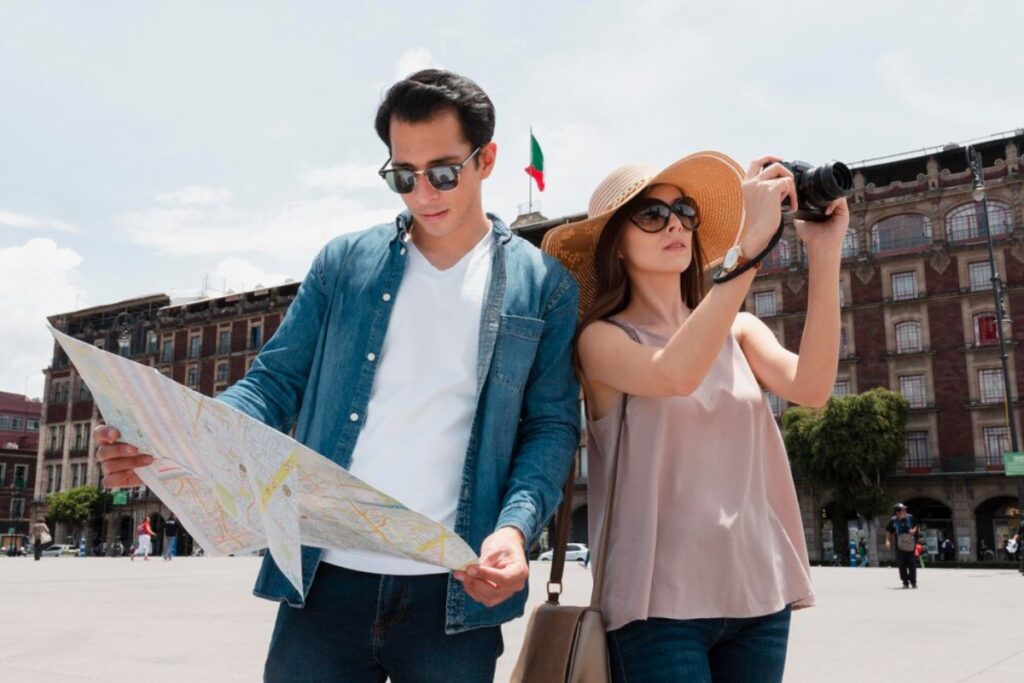 A couple stands in a city plaza, one holding a map while the other takes a photo, with historic buildings in the background and a flag.