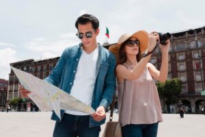 A couple stands in a city plaza, one holding a map while the other takes a photo, with historic buildings in the background and a flag.