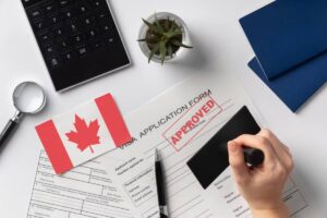 A hand stamps an APPROVED seal on a Canadian visa application, surrounded by a calculator, a pen, and a Canadian flag.