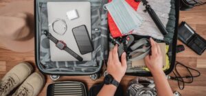 An open suitcase filled with travel essentials, including a laptop, smartphone, camera, masks, and a notebook, beside hiking shoes and a hat.
