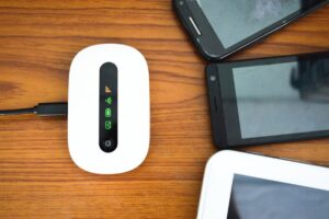 A white portable Wi-Fi router with LED indicators is connected by a cable, surrounded by three smartphones on a wooden table.