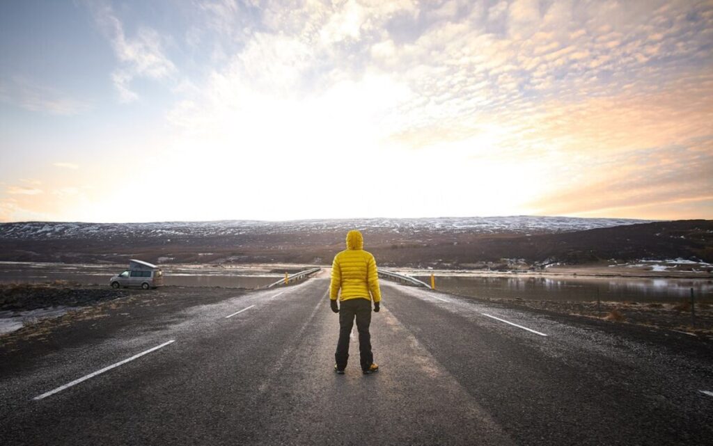 A person in a bright yellow jacket stands on an empty road overlooking a scenic landscape at sunset, with a van parked nearby.