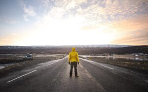 A person in a bright yellow jacket stands on an empty road overlooking a scenic landscape at sunset, with a van parked nearby.