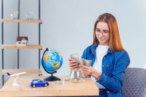 A person is counting cash in front of a globe, a toy car, a model airplane, and a glass jar filled with coins on a desk.