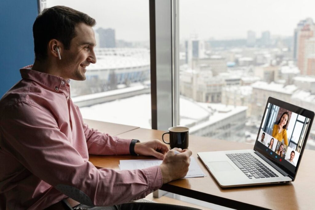 A person in a pink shirt sits at a desk with a laptop, participating in a video call, and holds a coffee cup with a city view outside.