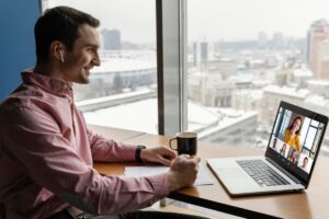 A person in a pink shirt sits at a desk with a laptop, participating in a video call, and holds a coffee cup with a city view outside.