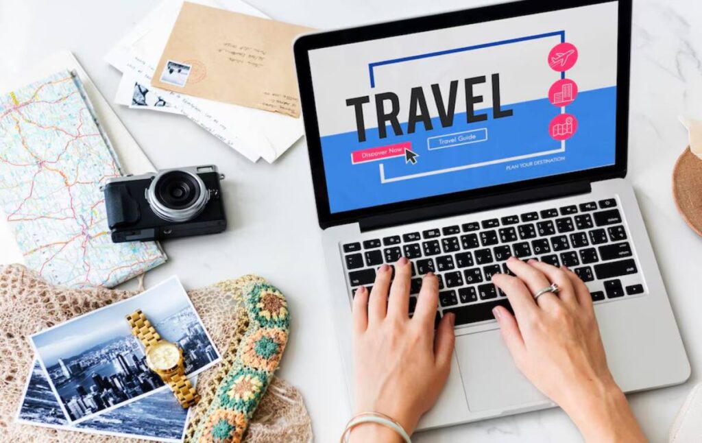 A person browses travel options on a laptop surrounded by a camera, map, postcards, and a watch, evoking wanderlust and exploration.