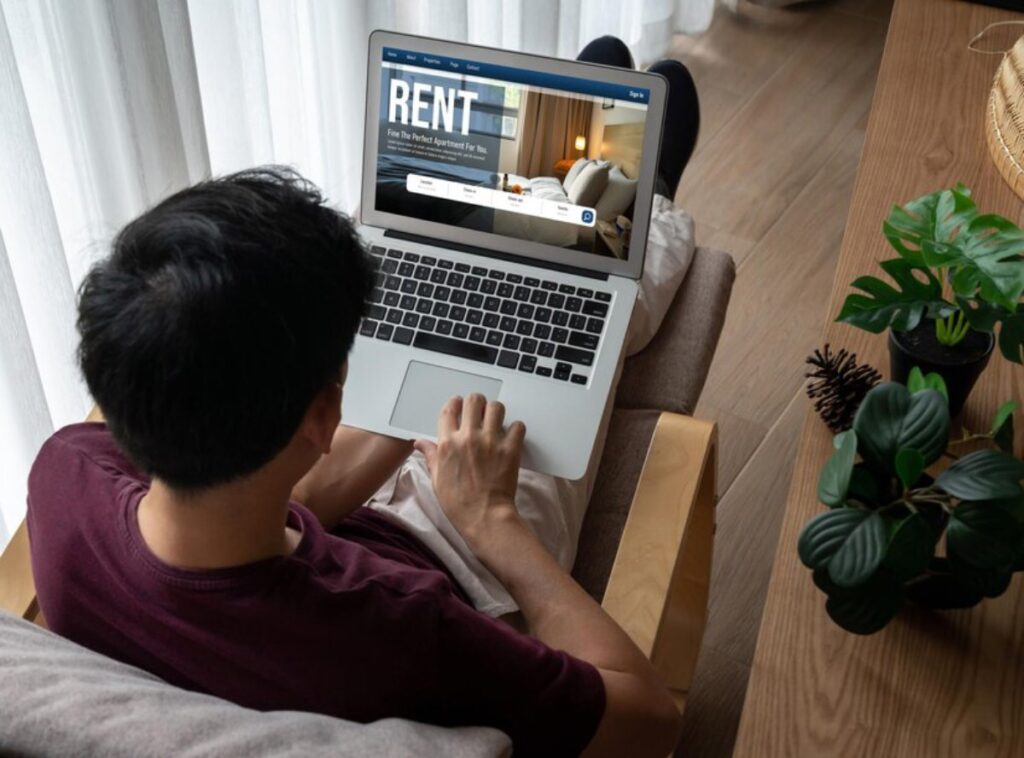 A man sits comfortably in an armchair, using a laptop to browse rent listings for apartments. Sunlight filters through sheer curtains.
