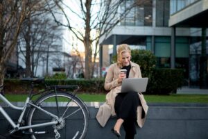 A woman in a beige trench coat sits on a stone ledge, sipping coffee while working on a laptop beside her bicycle in an urban setting.