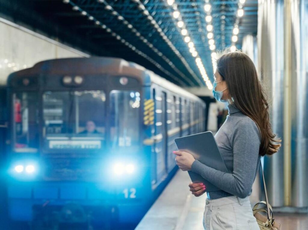 A woman stands at a train station holding a tablet, as a blue subway train approaches, illuminated by bright overhead lights.