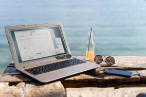 A laptop displaying a calendar sits on a rustic table by the beach, accompanied by sunglasses and a drink on a sunny day.