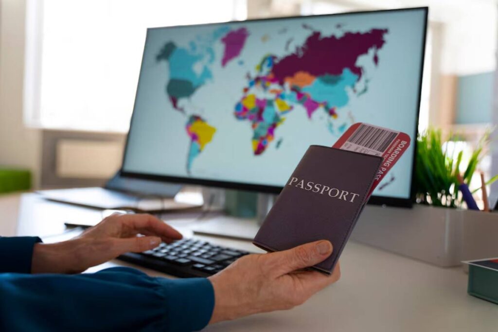 A person holds a passport and boarding pass in front of a computer displaying a colorful world map, symbolizing travel preparation.