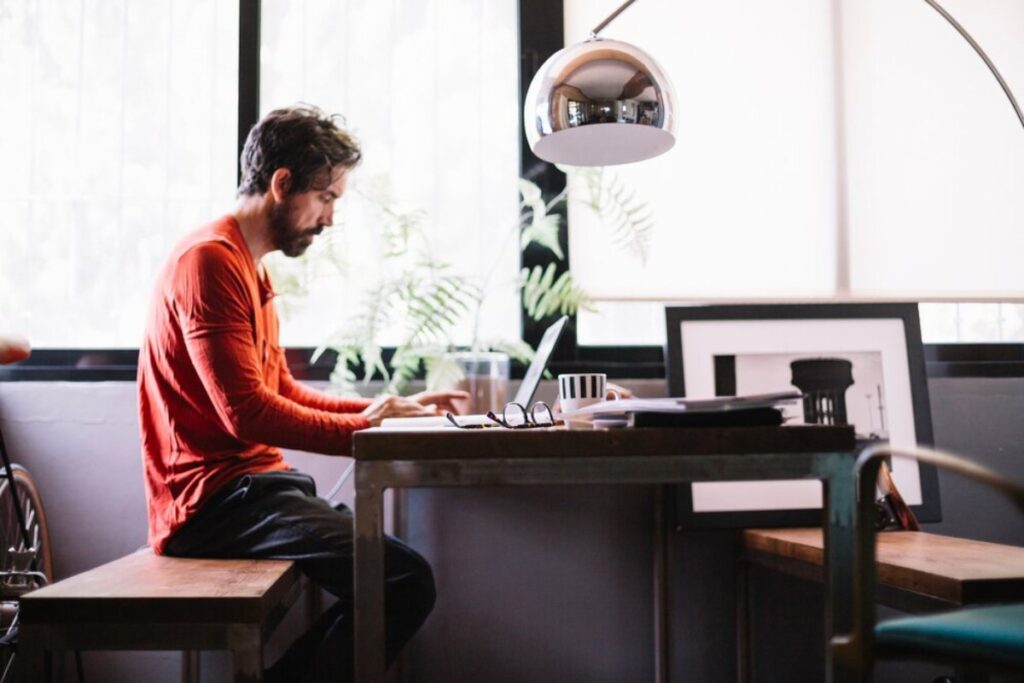 A person in an orange shirt sits at a wooden desk, working on a laptop in a bright, modern workspace with plants and framed pictures.
