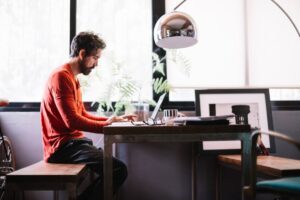 A person in an orange shirt sits at a wooden desk, working on a laptop in a bright, modern workspace with plants and framed pictures.