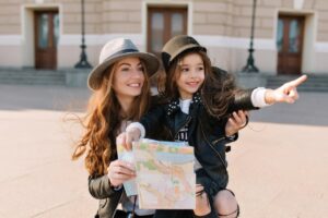 Two young women in stylish outfits hold a map, with one pointing excitedly while standing outdoors near a building.