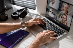 A person typing on a laptop displaying a video call, with headphones and a coffee cup nearby, on a well-lit desk.