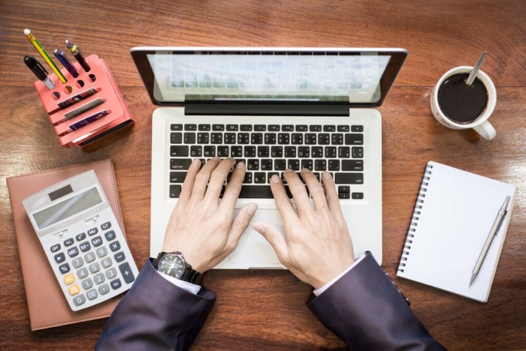 A person's hands type on a laptop keyboard, surrounded by a calculator, notebook, pen, coffee mug, and pencil holder on a wooden desk.