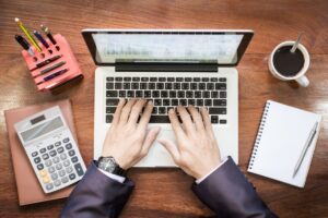A person's hands type on a laptop keyboard, surrounded by a calculator, notebook, pen, coffee mug, and pencil holder on a wooden desk.