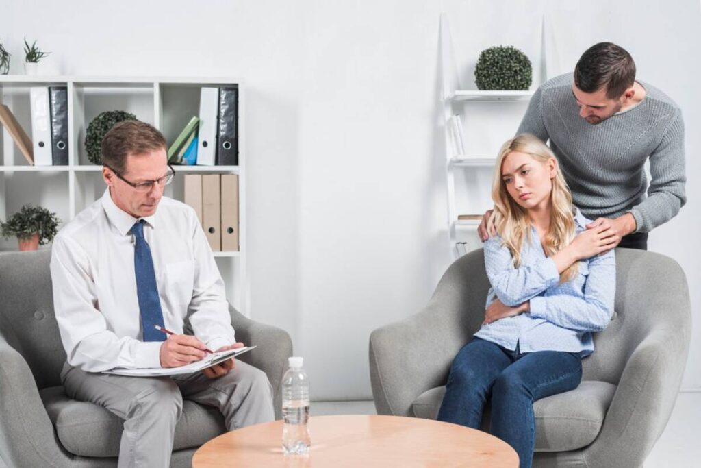 A therapist takes notes while a woman sits in a chair, comforted by a man who rests his hand on her shoulder in a modern office setting.