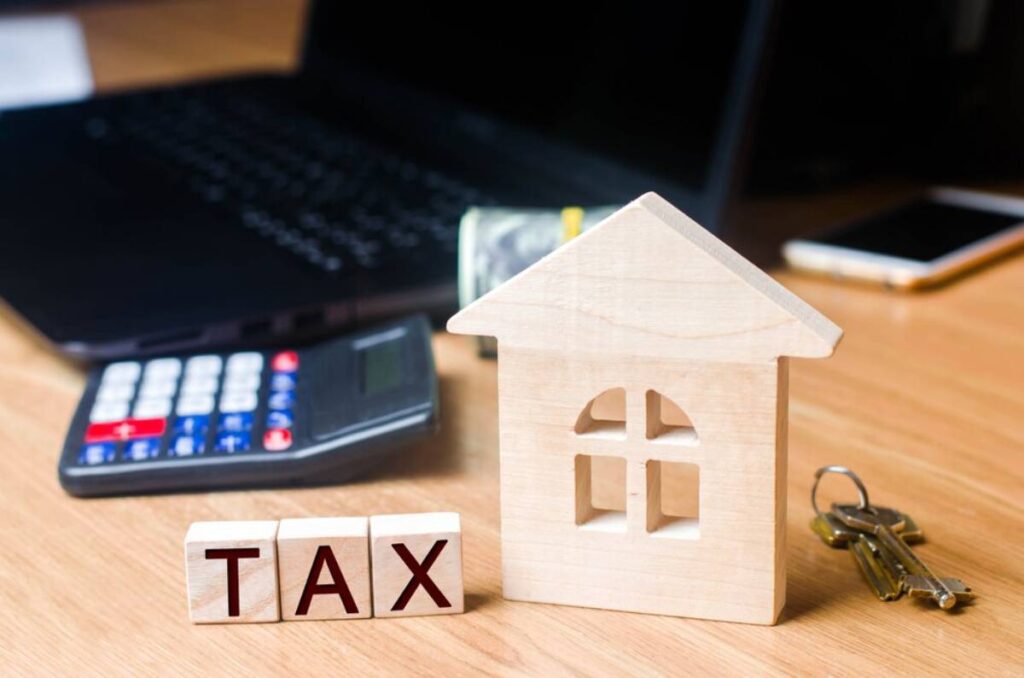 A wooden house model and TAX blocks next to a calculator and laptop, symbolizing real estate taxes and financial planning.