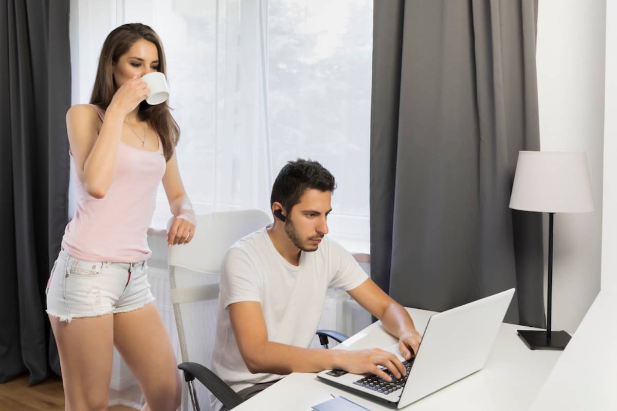 A woman in a pink tank top sips from a cup while standing next to a man using a laptop at a modern desk in a bright room.